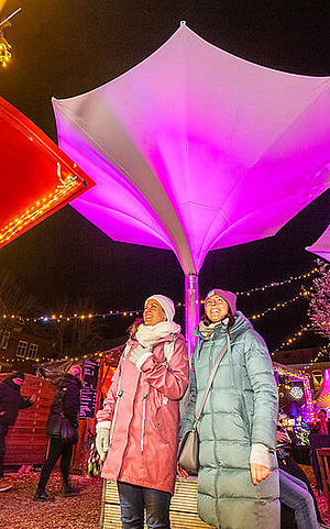 Eine Frau in einem pinken Mantel steht an einem liebevoll dekorierten Stand, während die Umgebung mit Lichtern und weihnachtlicher Dekoration einladend gestaltet ist. 