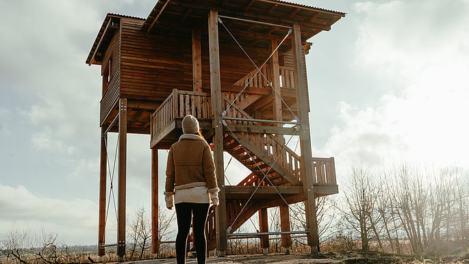 Ein Holzturm im Gegenlicht, davor steht eine Frau und schaut hinauf.