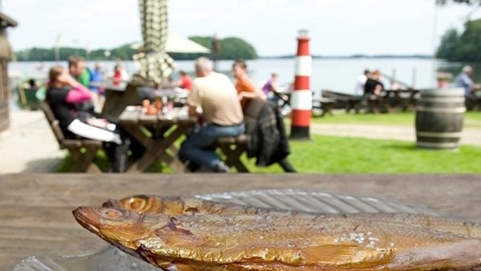 Geräucherter Fisch auf einem Teller. Im Hintergrund ist der See und eine Gruppe Menschen zu sehen.