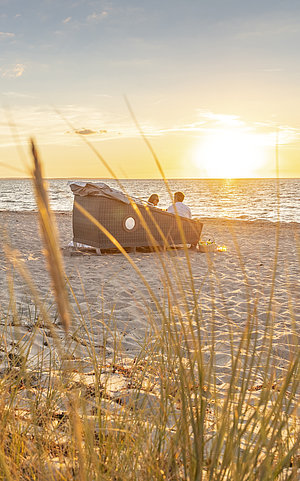 Ein offener Schlafstrandkorb im Gegenlicht am Strand, im Hintergrund glitzert das Meer im Abendlicht. 