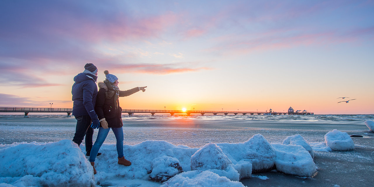 Ein Paar geht am Strand spazieren. Hinter ihnen ist die Ostsee zu sehen mit der Grömitzer Seebrücke. Am Strand liegen über all kleine weiße Schneehaufen. Das Paar hat dicke Winterjacken an, mit Schal und Mütze.