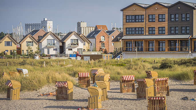 Im Vordergrund ist der Strand mit einzelnen Strandkörben, die unterschiedlich ausgerichtet, zu sehen. Im Hintergrund sind Hotels und Ferienwohnung von Heilgenhafen zu sehen.